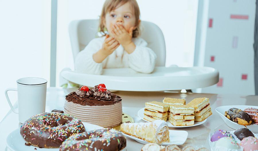Imagen de una niña pequeña sentada en una silla alta enfrente de una variedad de tortas y postres ubicados sobre una mesa.