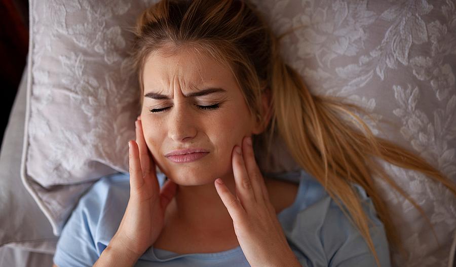 Una fotografía muestra a una mujer tocando su mandíbula con sus manos y apretando sus dientes mientras está recostada. Su rostro tiene una expresión de dolor.